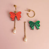 A pair of mismatched earrings on a pink background: one features a red bow, gold bar, and freshwater pearl; the other showcases a green bow with a gold-plated c-hoop, gold bar, and pearl for an interchangeable dangle earrings look.