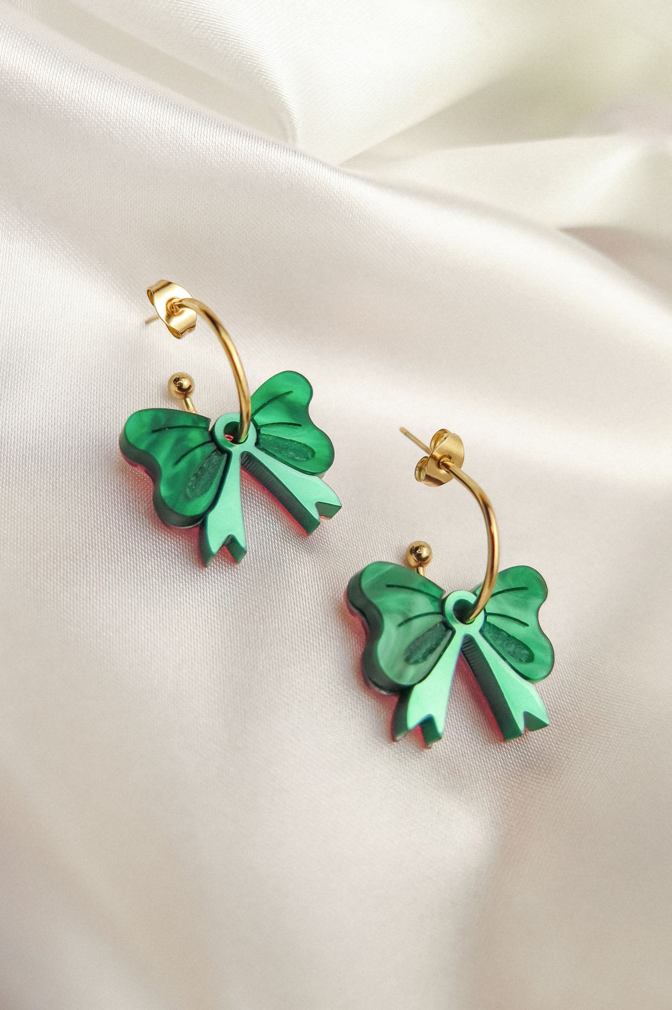 A pair of gold-plated c-hoop earrings with green bow-shaped charms and delicate freshwater pearl accents rests on soft, ivory satin fabric.