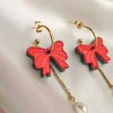 A pair of gold-plated c-hoop earrings with red butterfly-shaped charms and dangling gold chains ending in a single freshwater pearl, displayed on white satin fabric.