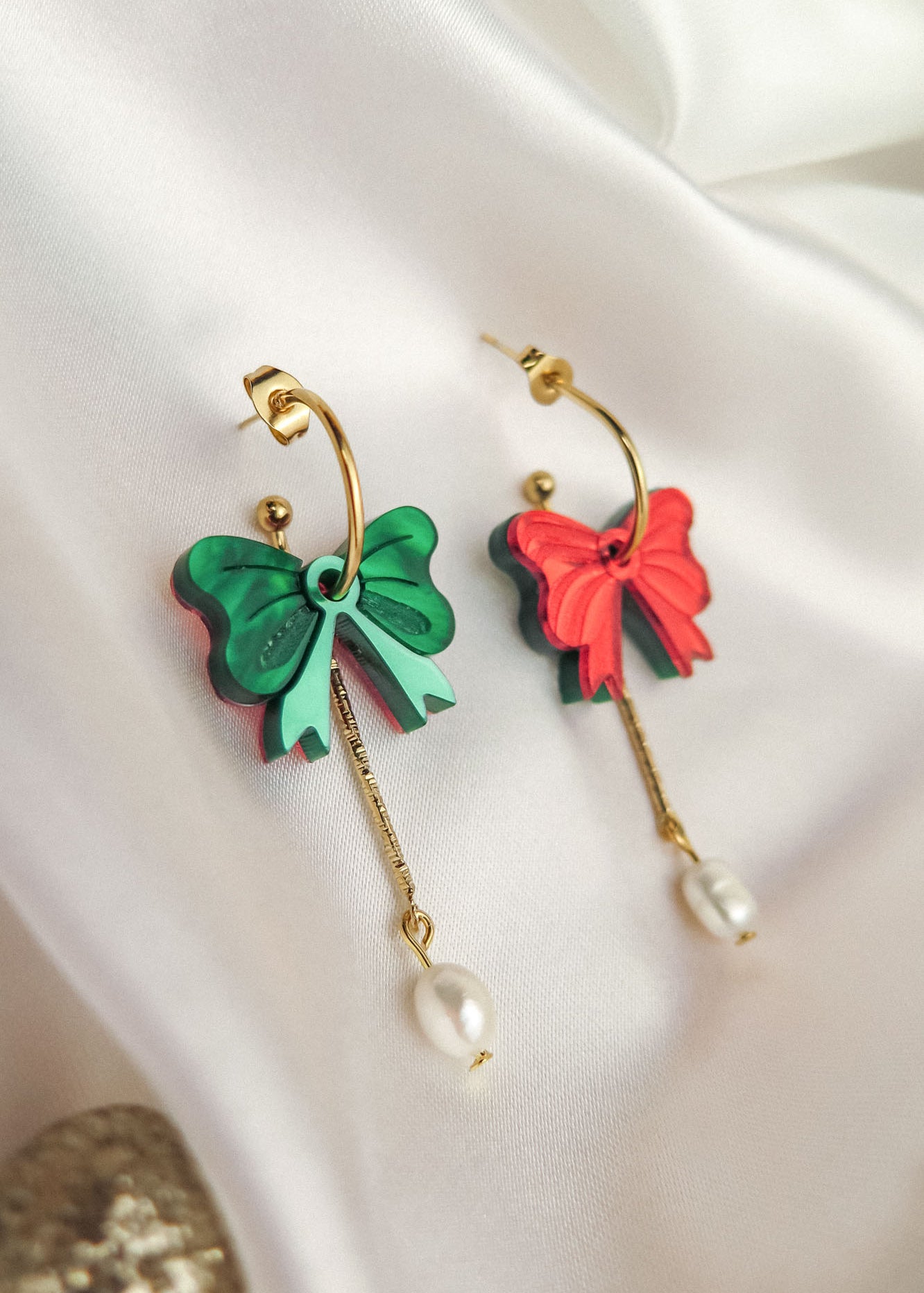 Two gold-plated c-hoop earrings with bow-shaped charms, one green and one red, each featuring a freshwater pearl dangling from a gold chain—stylish interchangeable dangle earrings displayed on white fabric.