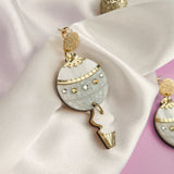 A decorative festive earring with a round, silver and gold design, studded with gems, rests on white satin fabric with gold and glitter accents. The perfect holiday jewelry piece, set against a soft lavender background.