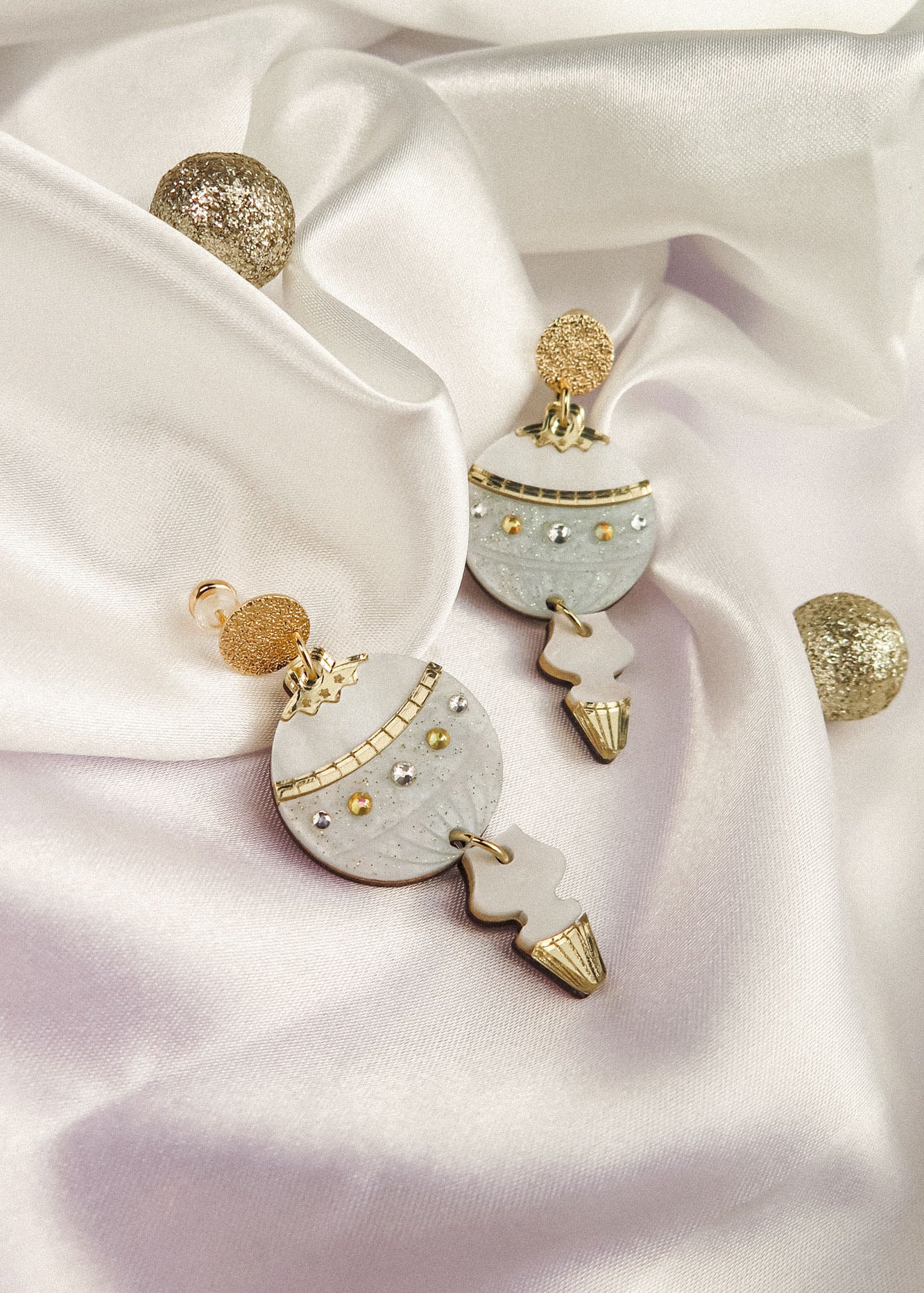 A pair of decorative Poinsettia Statement Hoops with gold, white, and silver details are displayed on white satin fabric, accompanied by glittery gold spheres—perfect as festive earrings to elevate your holiday jewelry collection.