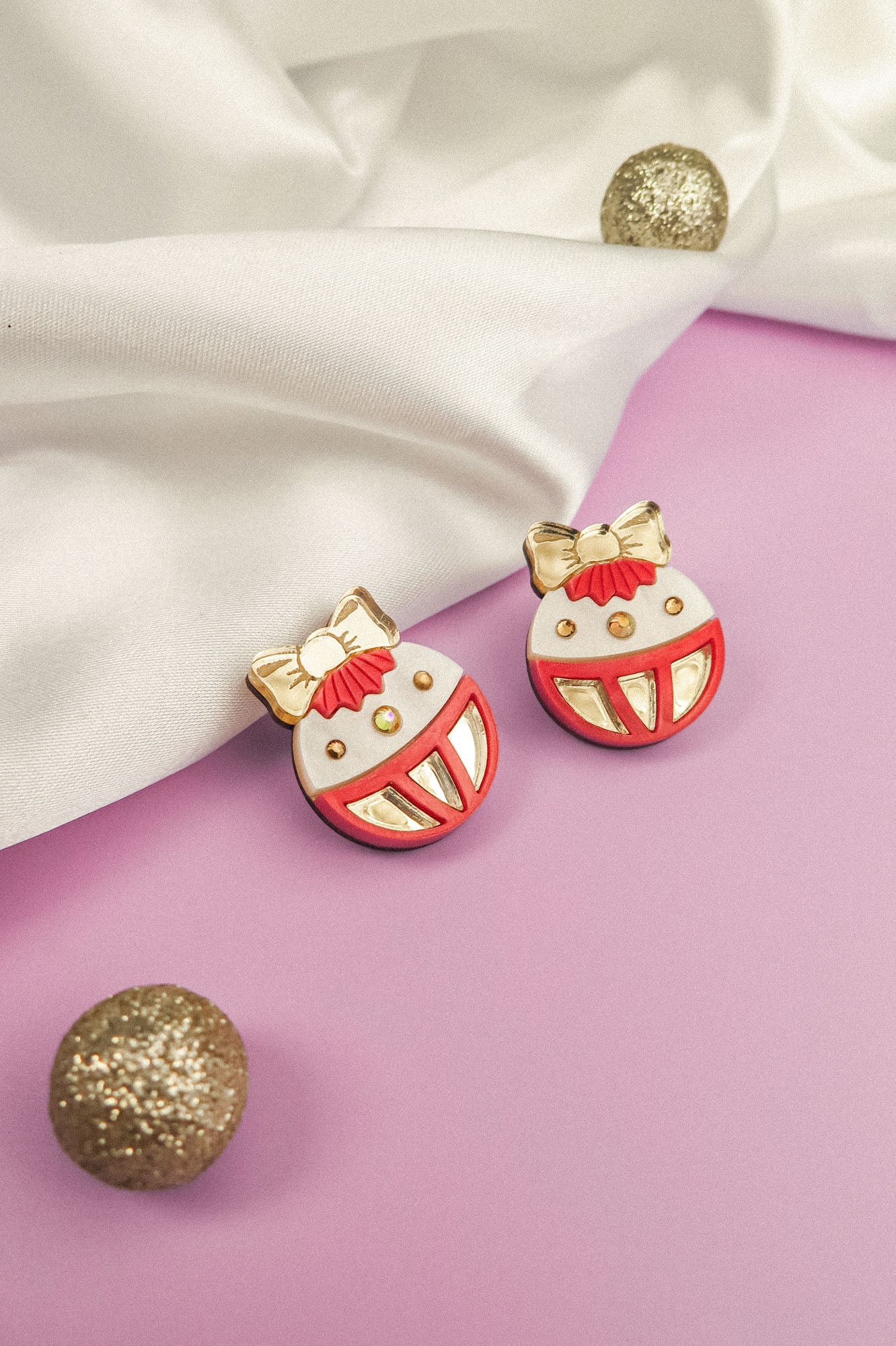 Two round festive earrings with red, white, and gold details shaped like ornaments with bows are displayed on a light purple surface next to a white cloth and glittery decorative balls—perfect holiday jewelry for any occasion.