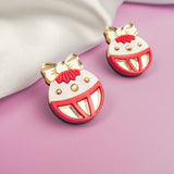 A pair of colorful enamel pins shaped like smiling faces with big red mouths, gold bows, and gold accents, bring a touch of festive earrings style, placed on a purple surface next to white fabric.