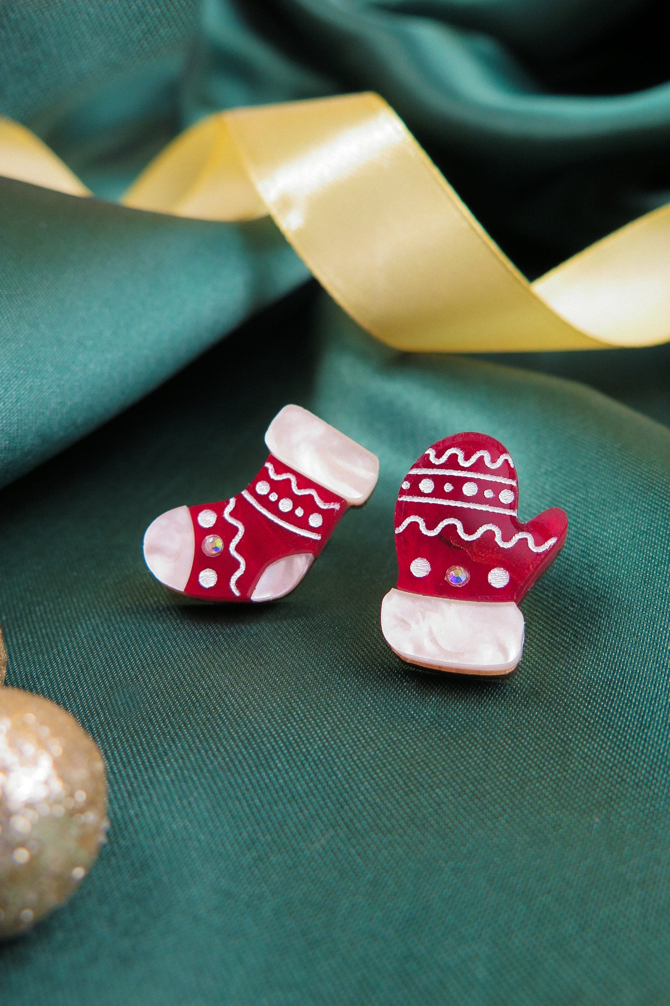 A festive holiday earrings set featuring Christmas Knitted Studs—a red mitten and red sock with white details and rhinestones—displayed on green fabric with a gold satin ribbon in the background.