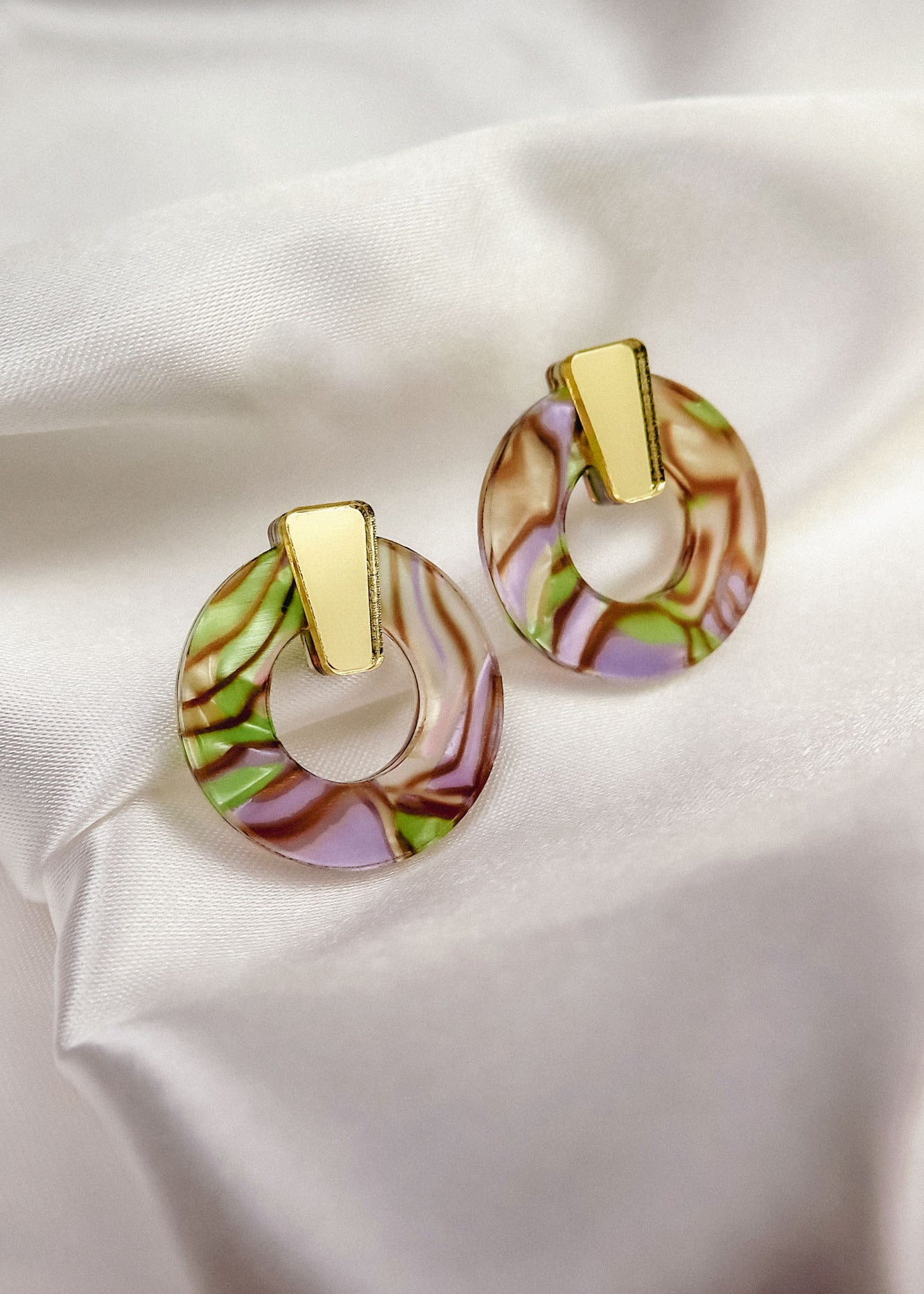 A pair of hypoallergenic Minimalist Circle Stud Earrings with gold rectangular tops and marbled acrylic glass circular bottoms in pink, green, and brown, displayed on white satin fabric.