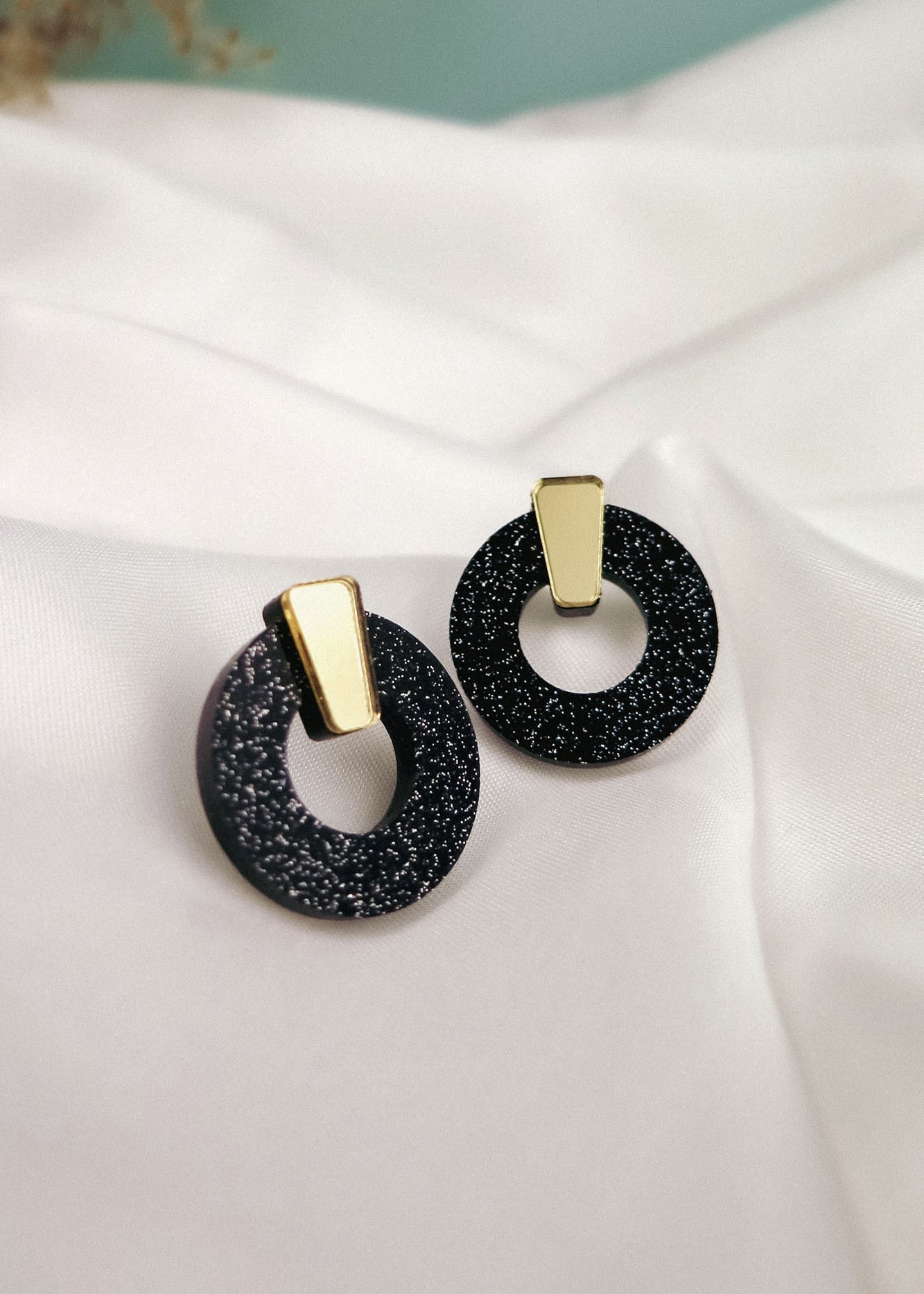 A pair of stylish Minimalist Circle Stud Earrings featuring black glittery acrylic glass hoops with gold rectangular accents, displayed on a white satin fabric background. Hypoallergenic for comfortable, all-day wear.
