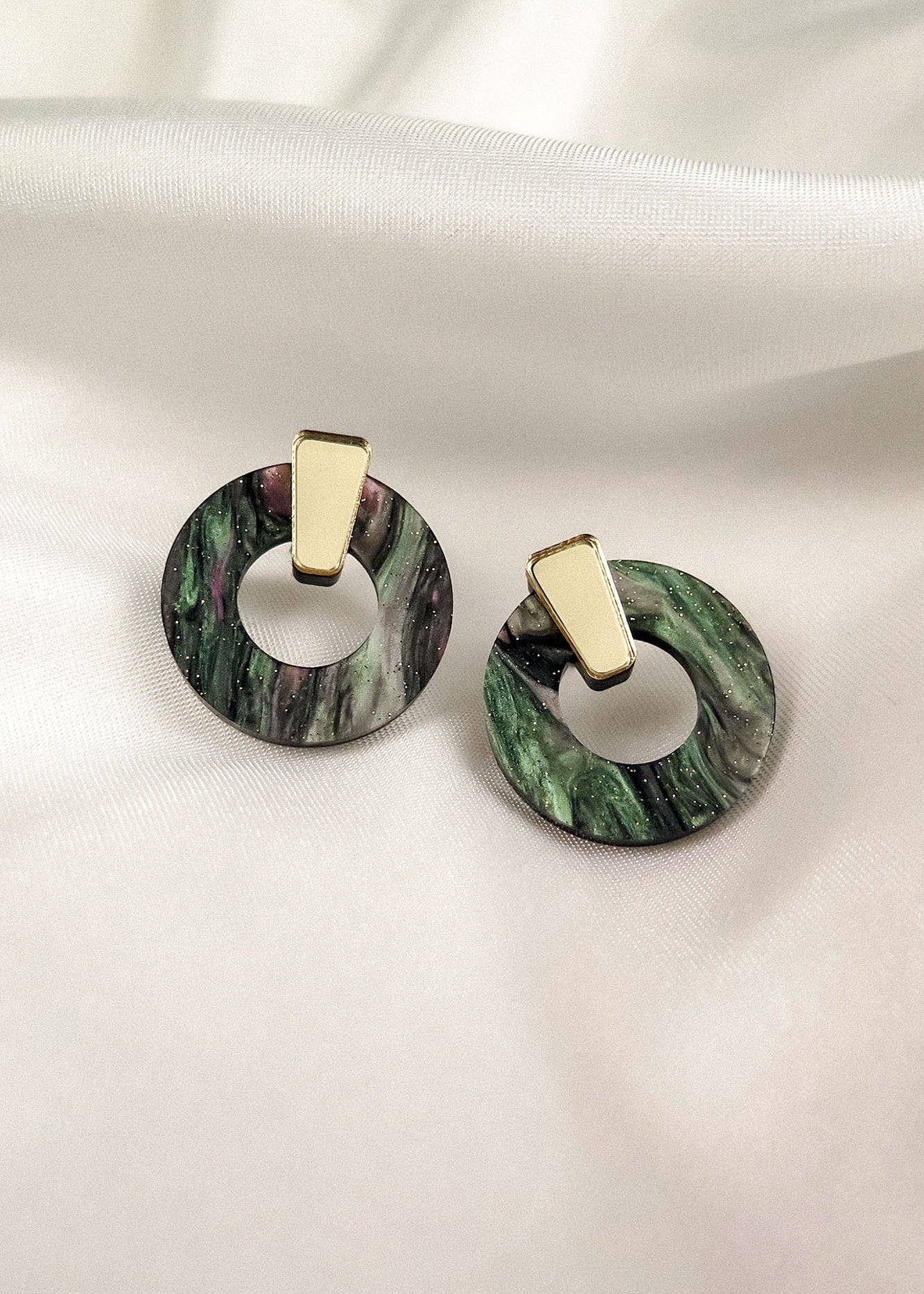 Minimalist Circle Stud Earrings featuring gold geometric studs and circular, marbled green and black hoops crafted from acrylic glass, displayed on a white fabric background. Hypoallergenic for comfortable wear.