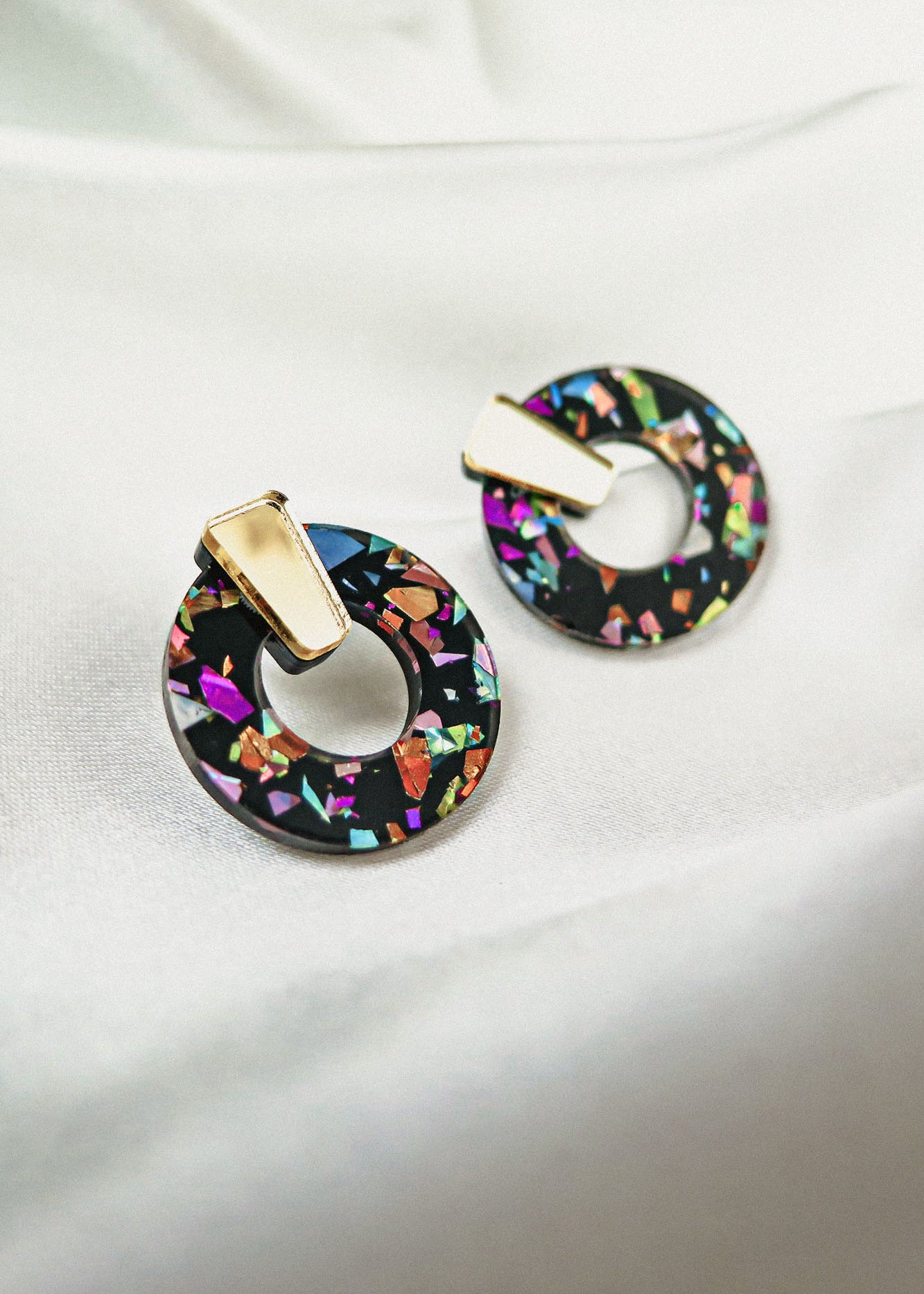 A pair of Minimalist Circle Stud Earrings made from hypoallergenic acrylic glass, featuring a black base with colorful, iridescent shards and gold-toned rectangular studs, displayed on a white fabric background.