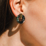 Close-up of a woman's ear wearing minimalist circle stud earrings with a geometric design, featuring a circular black disk and a gold rectangular piece. Her brown hair is tucked behind her ear; these are elegant hypoallergenic earrings.