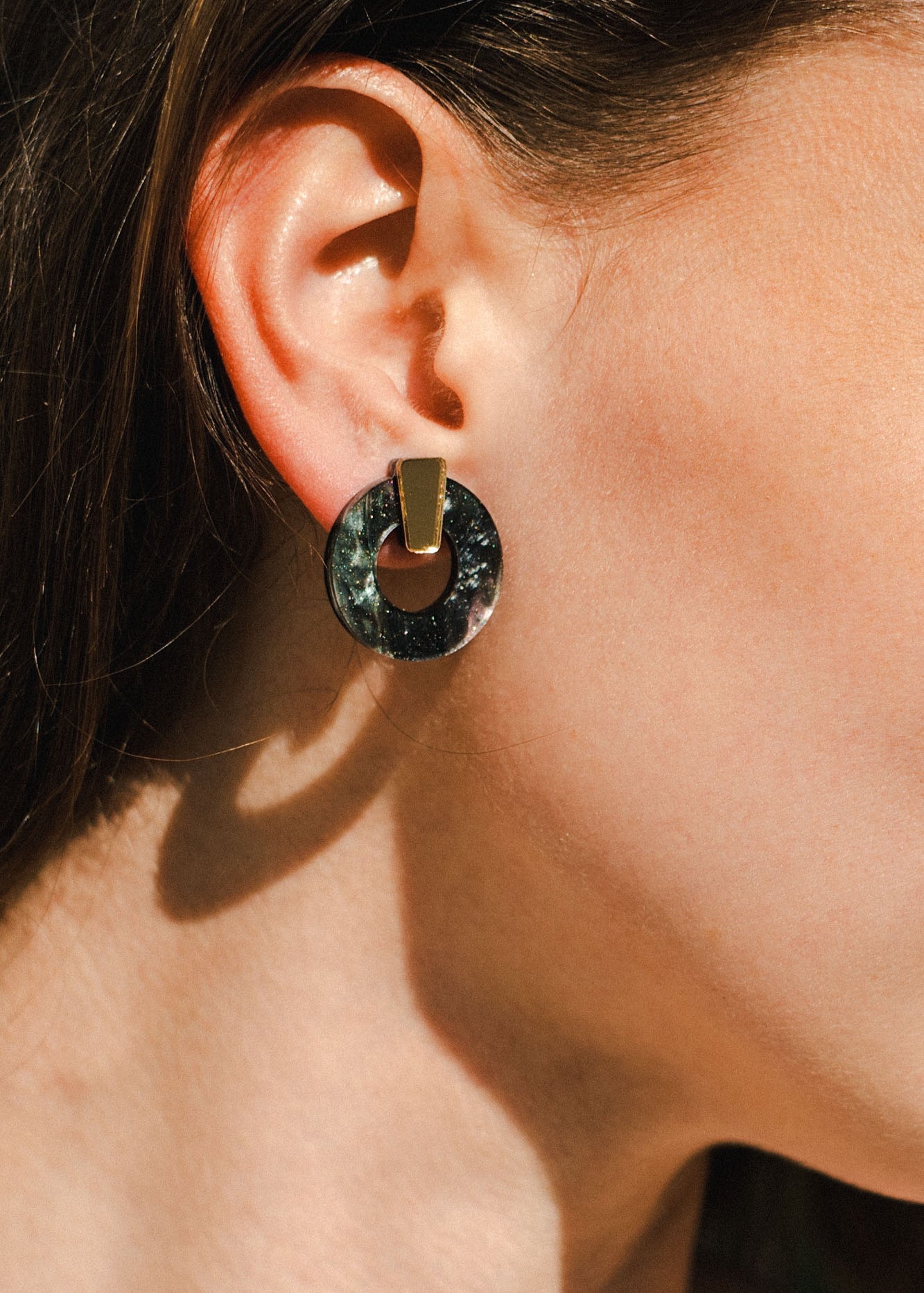 Close-up of a woman's ear wearing minimalist circle stud earrings with a geometric design, featuring a circular black disk and a gold rectangular piece. Her brown hair is tucked behind her ear; these are elegant hypoallergenic earrings.