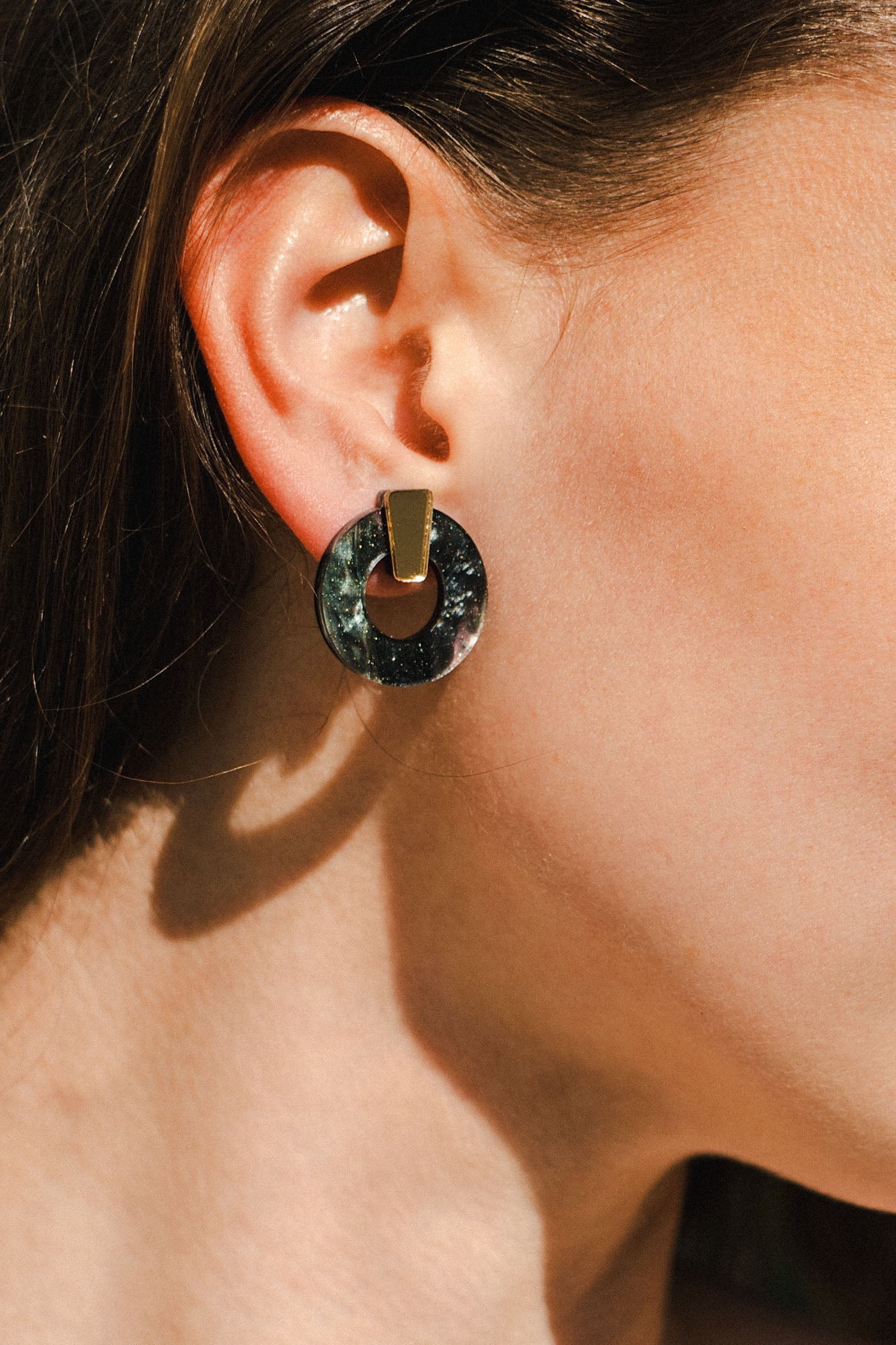 Close-up of a woman's ear wearing minimalist circle stud earrings with a geometric design, featuring a circular black disk and a gold rectangular piece. Her brown hair is tucked behind her ear; these are elegant hypoallergenic earrings.