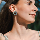 A woman with long brown hair and a tattoo on her arm poses outdoors in sunlight, wearing a green satin top and minimalist circle stud earrings, looking off to the side and smiling slightly.