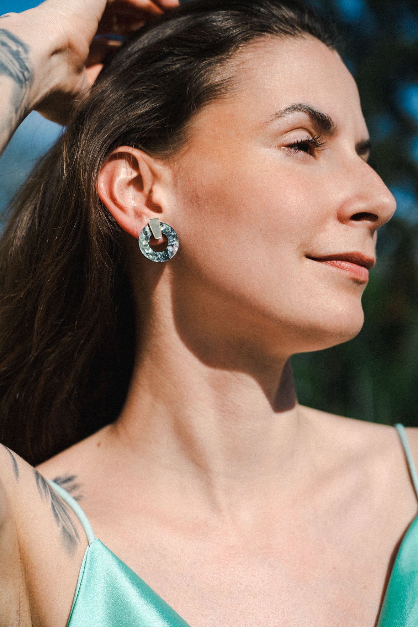 A woman with long brown hair and a tattoo on her arm poses outdoors in sunlight, wearing a green satin top and minimalist circle stud earrings, looking off to the side and smiling slightly.