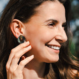 A woman with long brown hair smiles outdoors, holding her hand near her face to show off a large, round Minimalist Circle Stud Earring. The background is softly blurred with hints of greenery and blue sky.