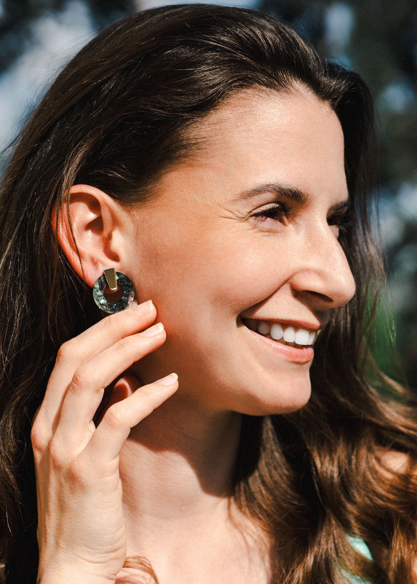 A woman with long brown hair smiles outdoors, holding her hand near her face to show off a large, round Minimalist Circle Stud Earring. The background is softly blurred with hints of greenery and blue sky.