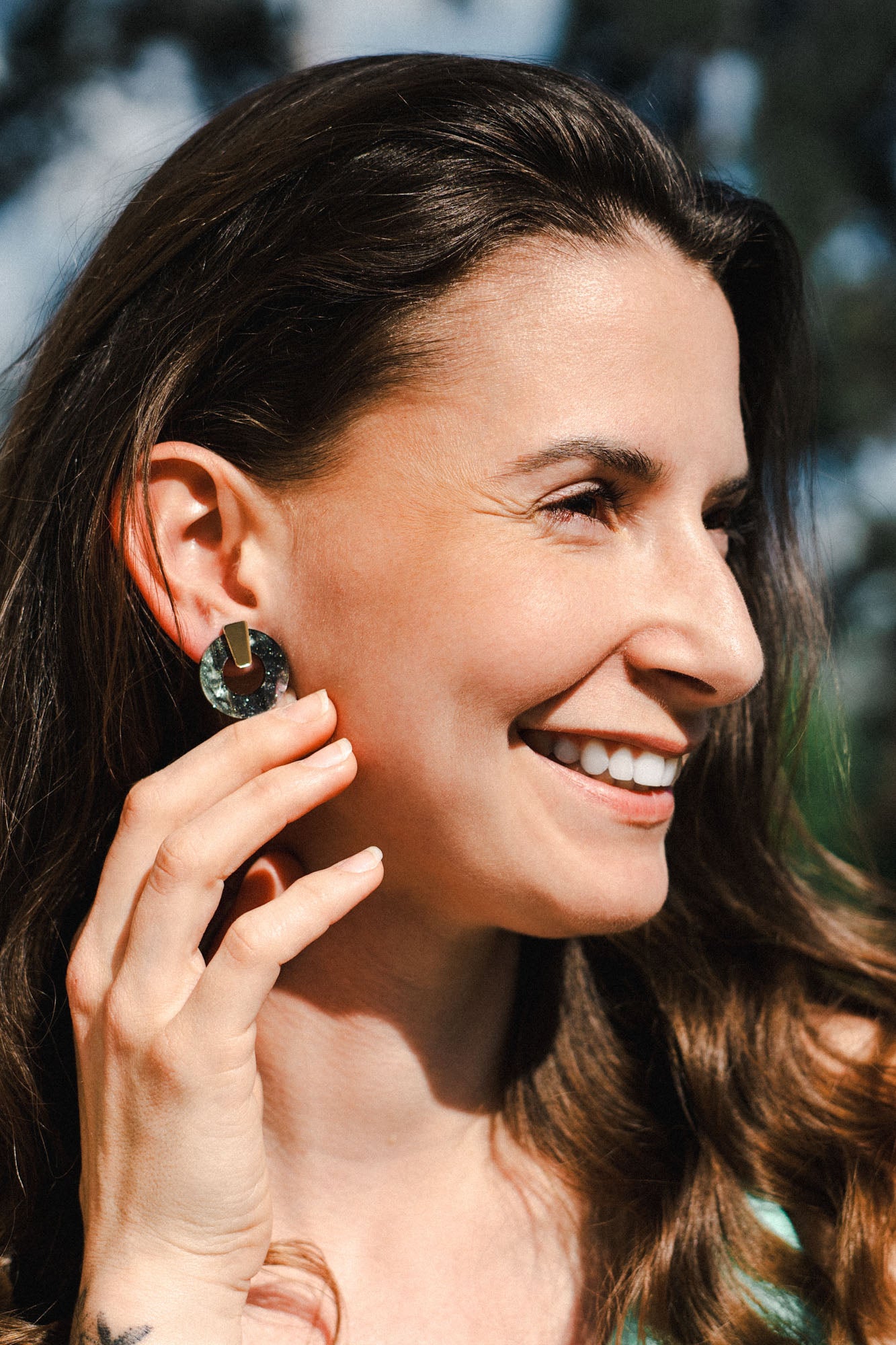 A woman with long brown hair smiles outdoors, holding her hand near her face to show off a large, round Minimalist Circle Stud Earring. The background is softly blurred with hints of greenery and blue sky.
