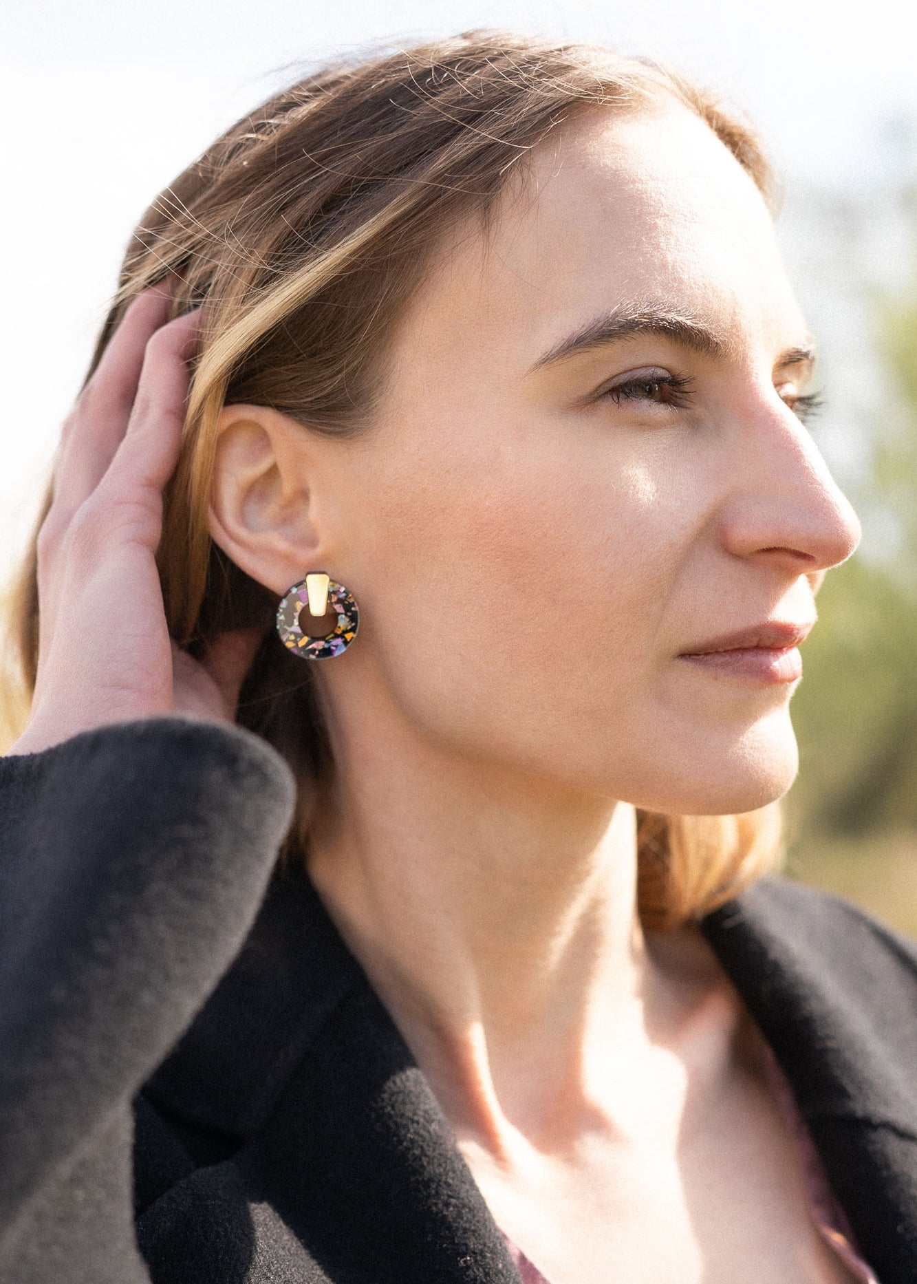 A woman with light brown hair touches her head while looking into the distance. She wears a black coat, a mauve top, and Minimalist Circle Stud Earrings that are hypoallergenic. The background is outdoors and softly blurred by sunlight.
