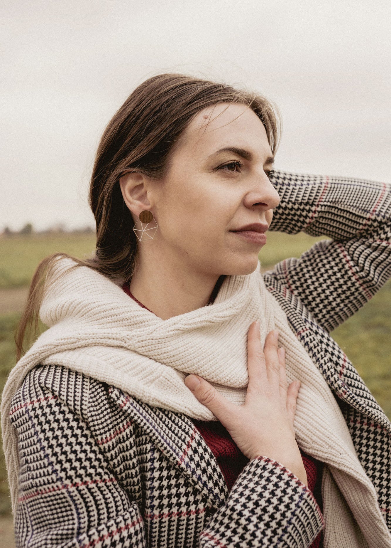 A woman in a houndstooth coat and cream scarf, embodying minimalist elegance, stands outdoors on a grassy, overcast day. She gazes thoughtfully into the distance, one hand on her chest and the other behind her head.
