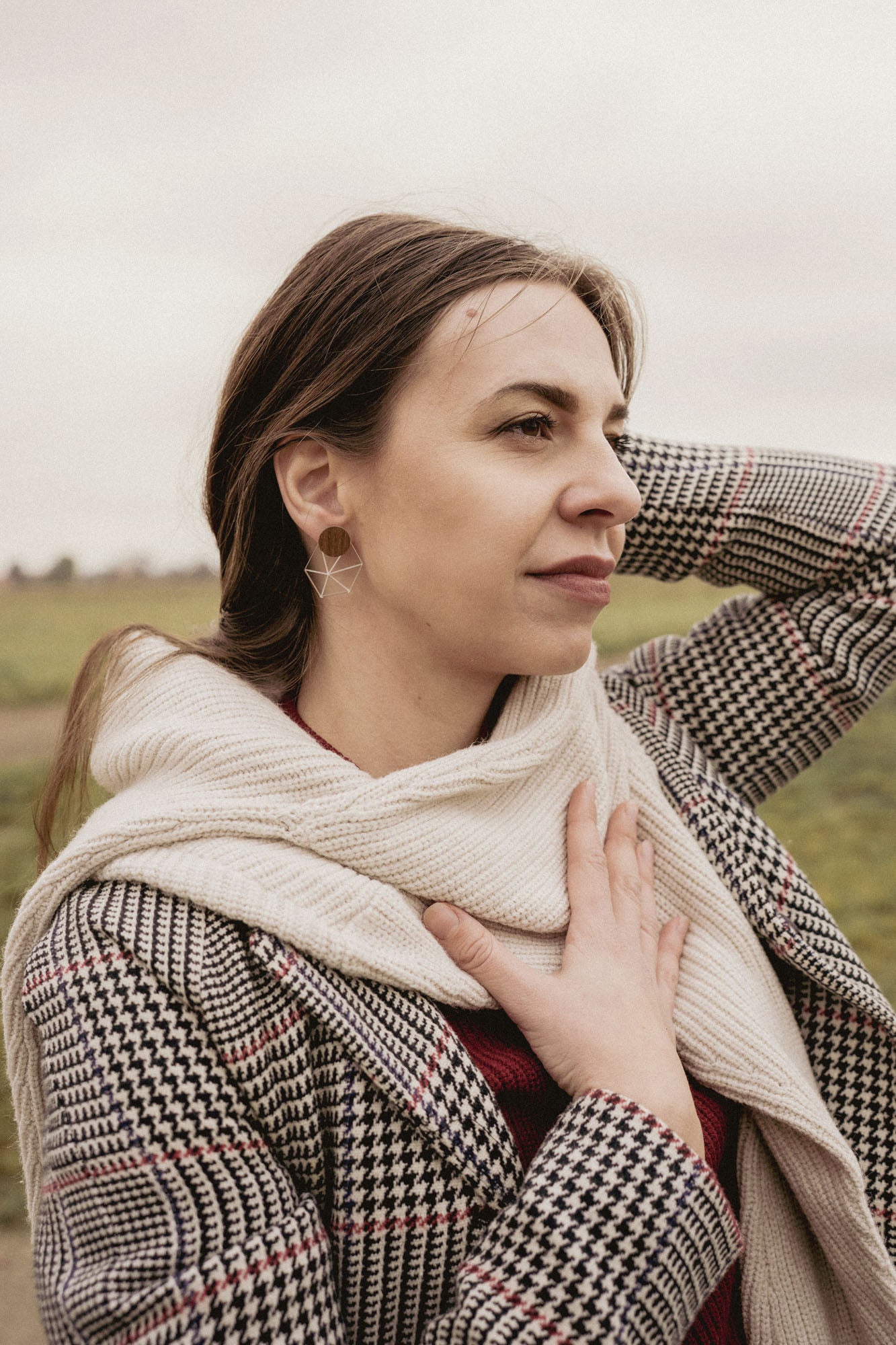 A woman in a houndstooth coat and cream scarf, embodying minimalist elegance, stands outdoors on a grassy, overcast day. She gazes thoughtfully into the distance, one hand on her chest and the other behind her head.