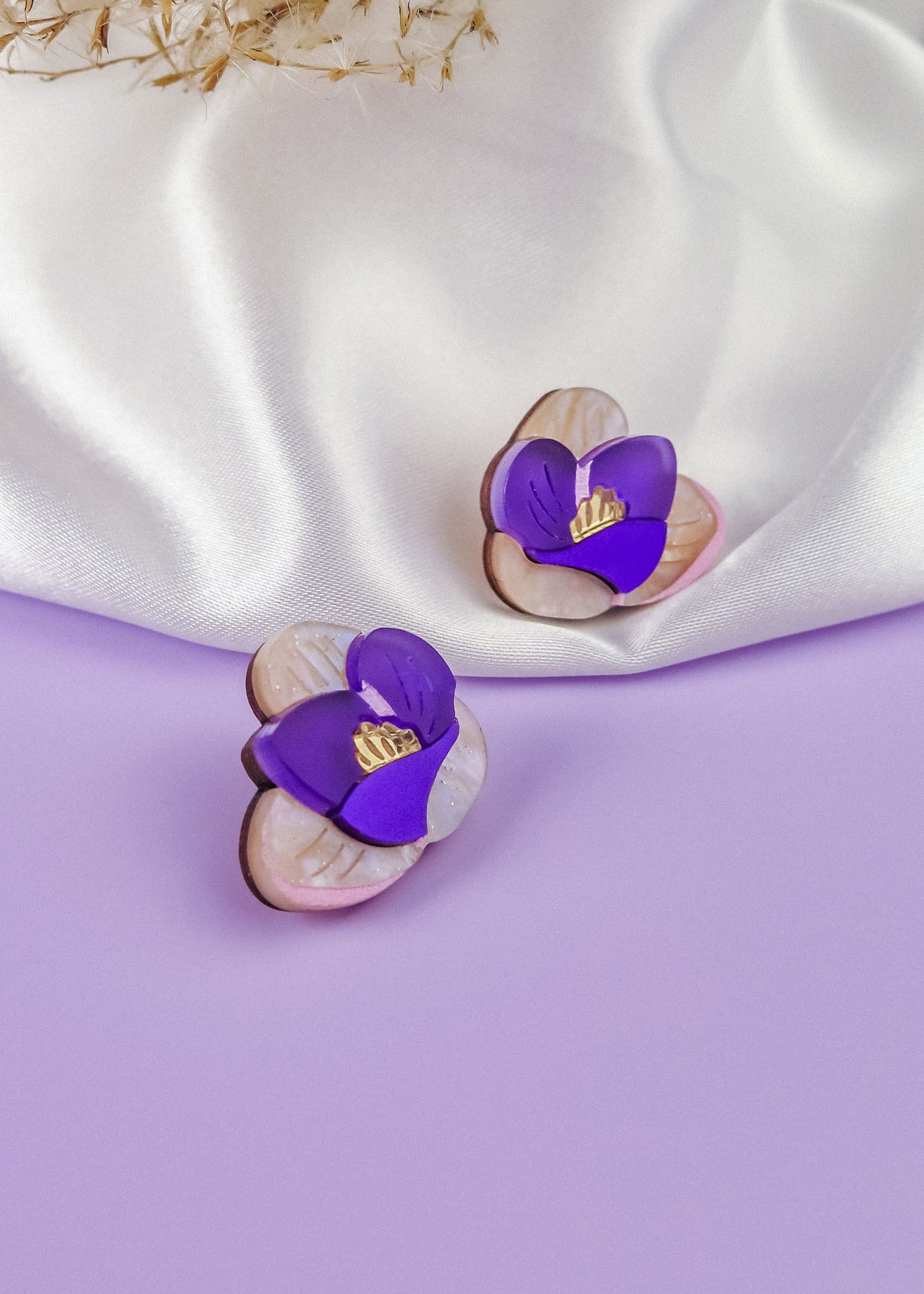 Two purple and white Crocus Stud Earrings, featuring hand-painted flowers, are displayed on a smooth lavender surface, with a white satin cloth and dried flowers in the background.