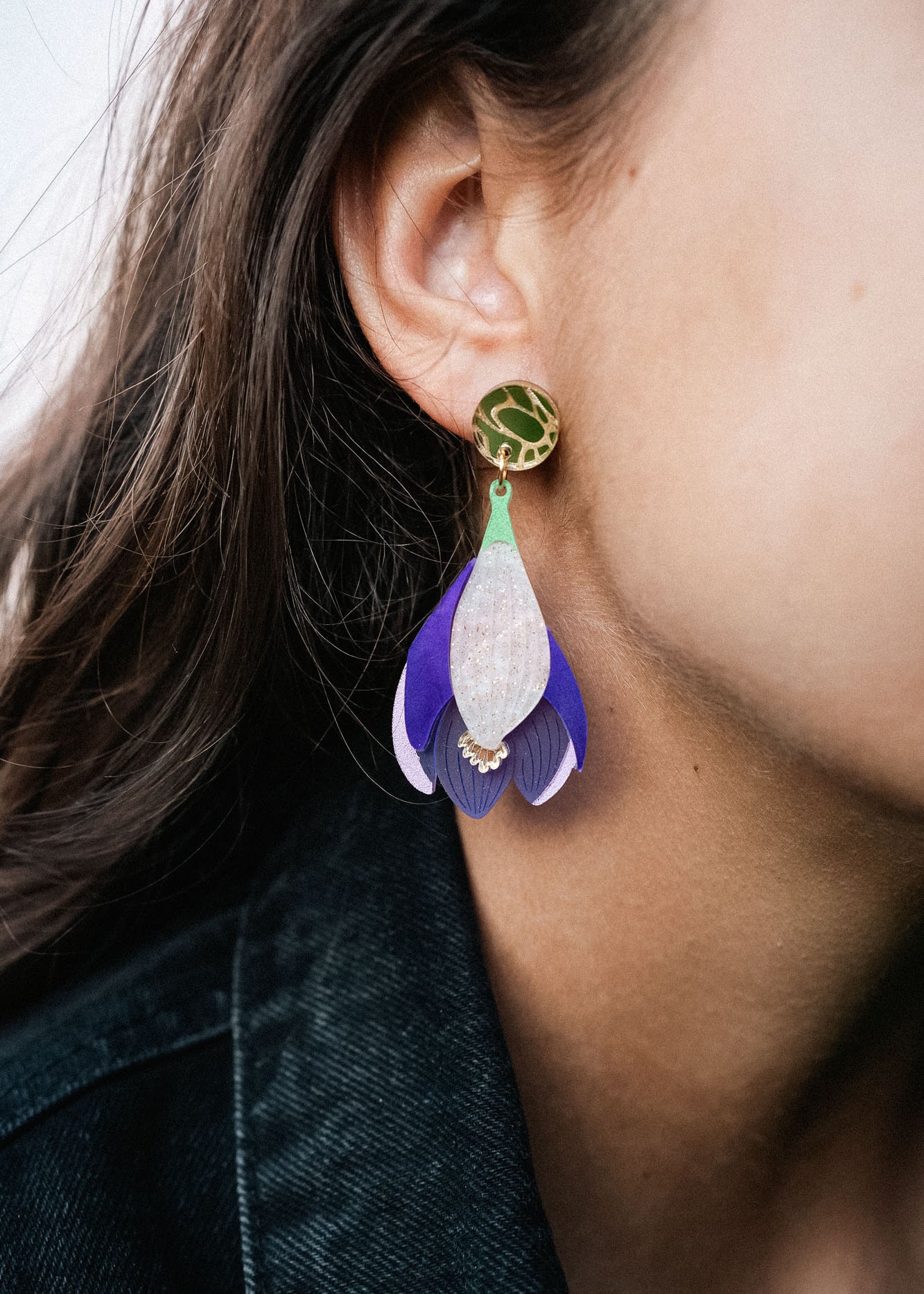 Close-up of a person wearing Crocus Flower Earrings—a handcrafted, statement accessory shaped like a flower petal in purple, white, and gold. The person has long brown hair and is dressed in a dark-colored top.