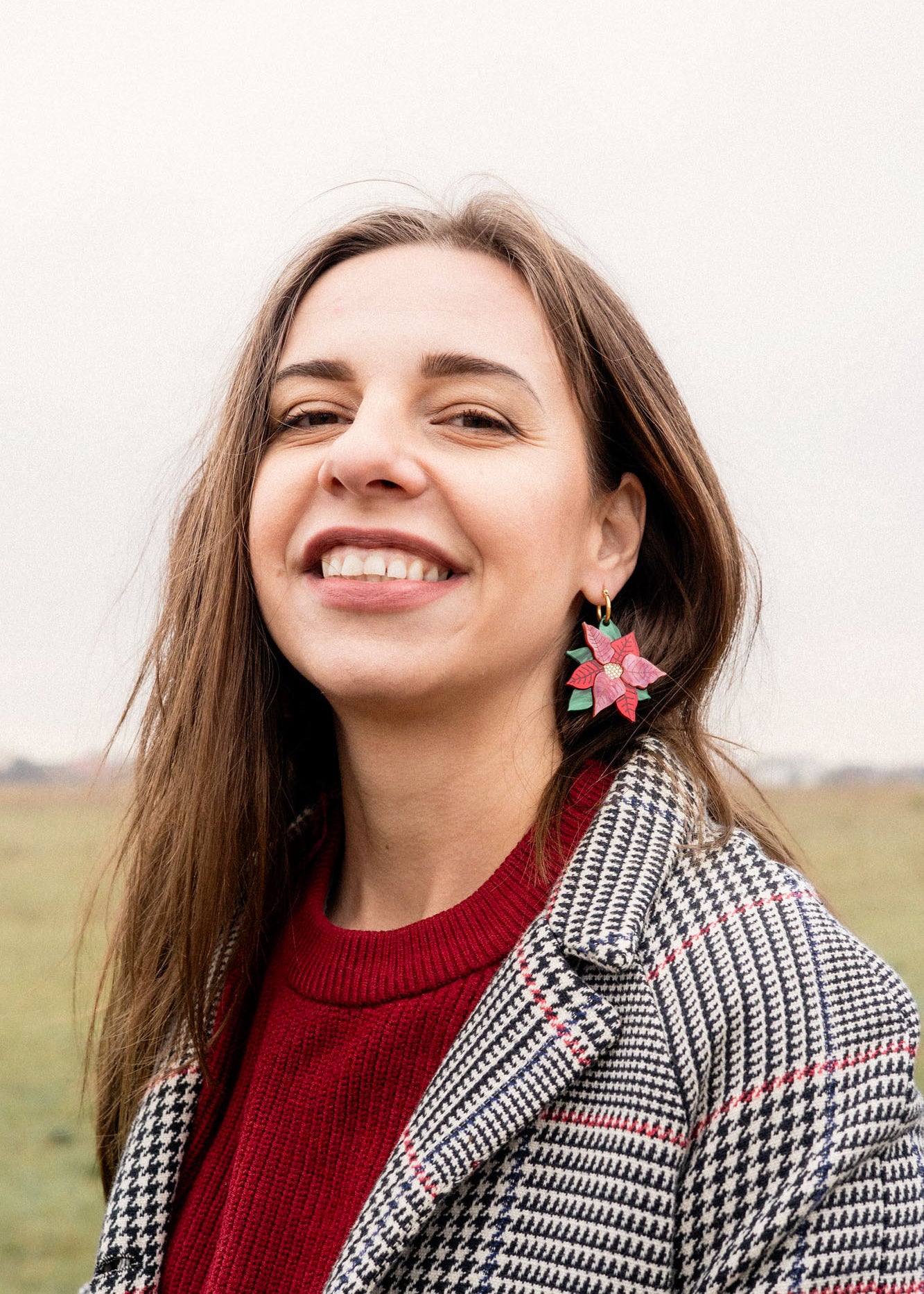 A woman with long brown hair smiles outdoors, wearing a red sweater, a black-and-white checkered coat, and festive holiday earrings. The background is a grassy field under an overcast sky.