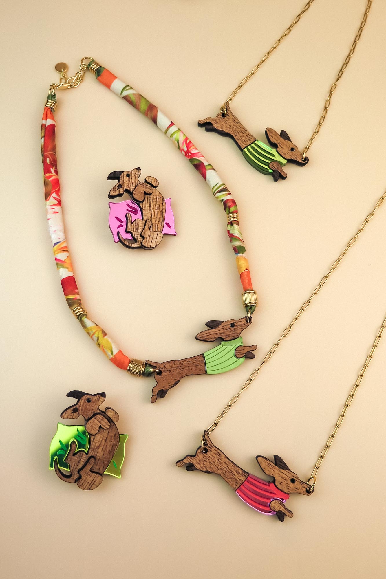 Colorful handcrafted jewelry featuring Dachshund necklaces and brooches with dogs in green and pink sweaters, set against a pale beige background. Each piece is thoughtfully made using recycled materials.