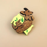 A playful brooch shaped like a kangaroo lies on a plain beige background. With engraved details and bright yellow and green leaf-like accents, this wooden piece adds whimsy to any dog accessory collection.