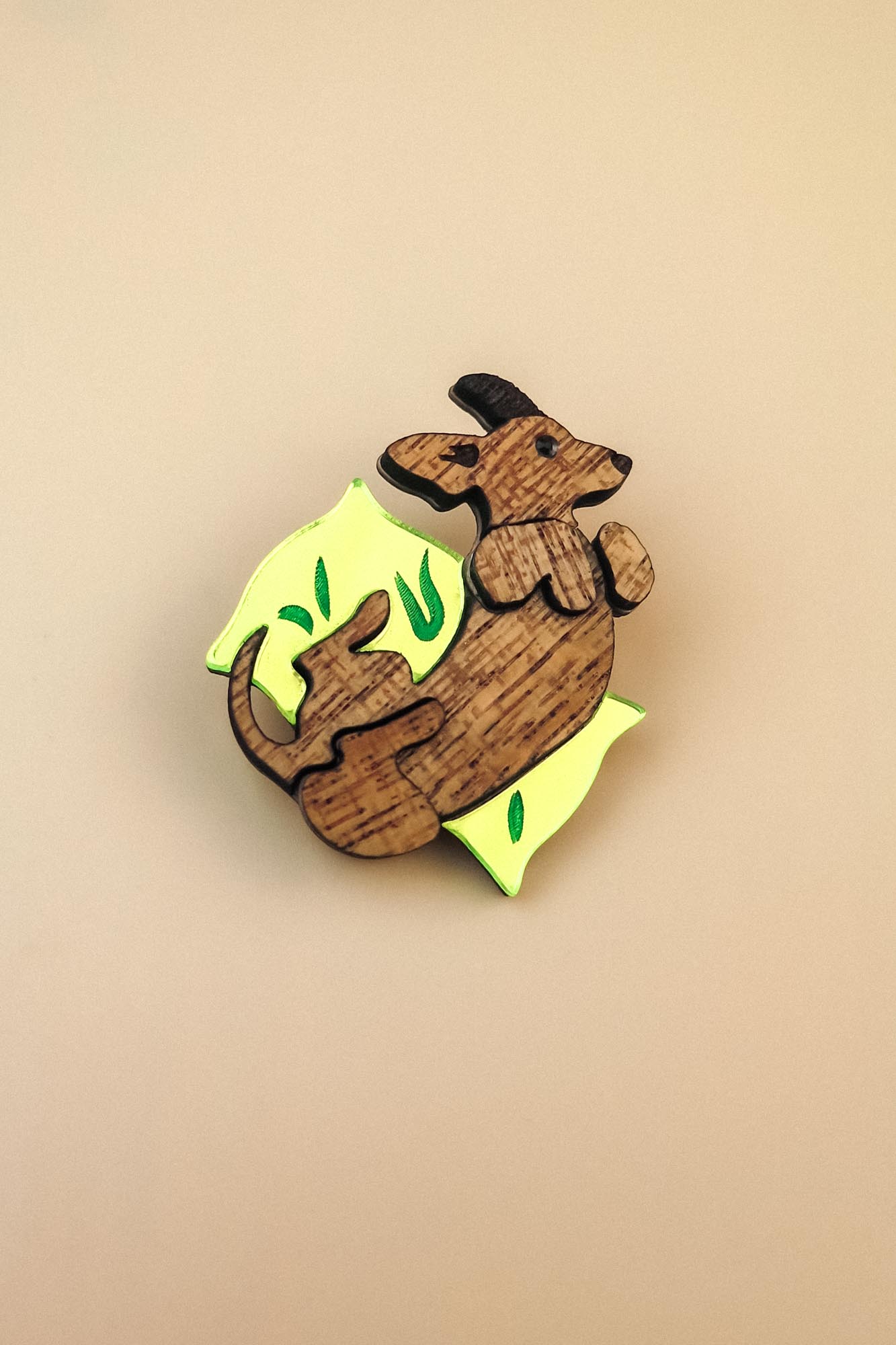 A playful brooch shaped like a kangaroo lies on a plain beige background. With engraved details and bright yellow and green leaf-like accents, this wooden piece adds whimsy to any dog accessory collection.