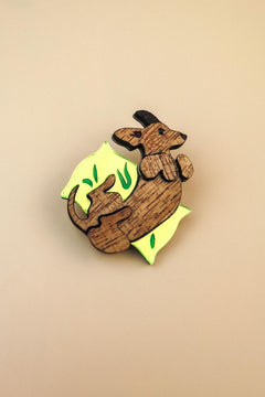 A playful brooch shaped like a kangaroo lies on a plain beige background. With engraved details and bright yellow and green leaf-like accents, this wooden piece adds whimsy to any dog accessory collection.
