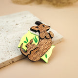 A playful brooch, this wooden pin shaped like a mouse lounging on a yellow wedge with green leaf accents rests on textured beige stone, making it a charming dog accessory or a unique Dachshund brooch for any outfit.