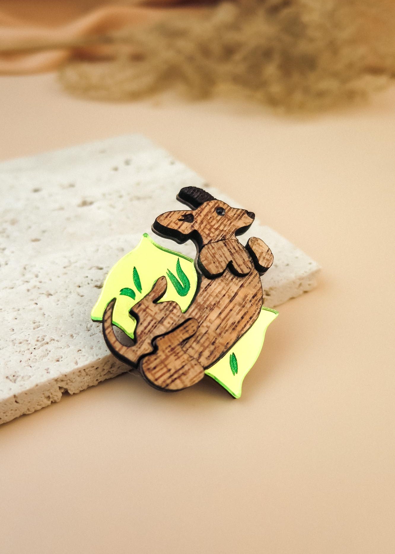 A playful brooch, this wooden pin shaped like a mouse lounging on a yellow wedge with green leaf accents rests on textured beige stone, making it a charming dog accessory or a unique Dachshund brooch for any outfit.