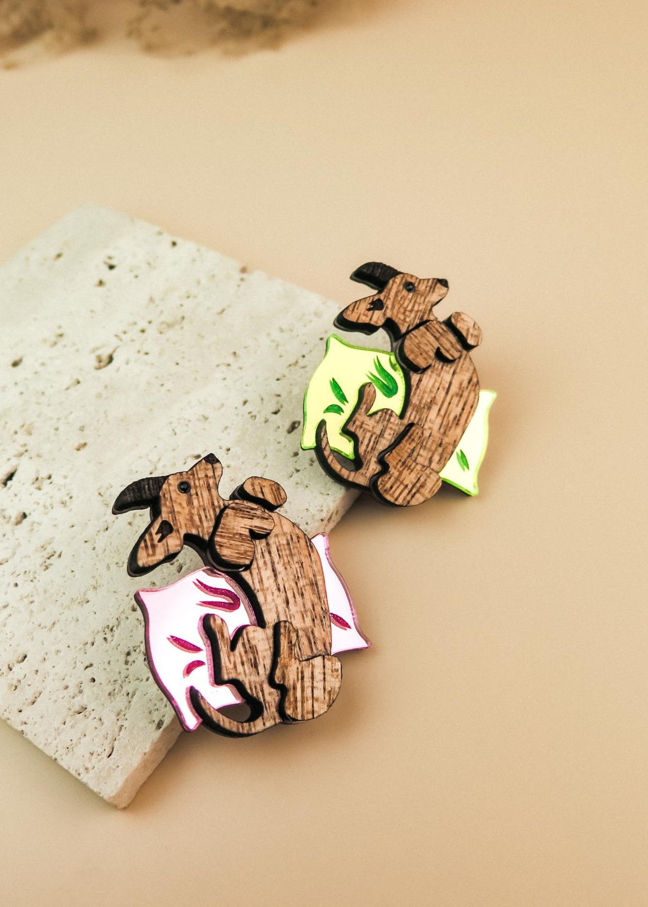 Two playful brooches shaped like reclining dachshunds, each resting on a colorful pillow—one pink and one neon green—placed on a textured stone surface. A charming touch for any dog accessory collection or fans of unique brooches.