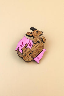 A wooden Dachshund brooch shaped like a brown dog lying on its back on a pink pillow with small decorative marks, set against a plain beige background—perfect for playful jewelry lovers.