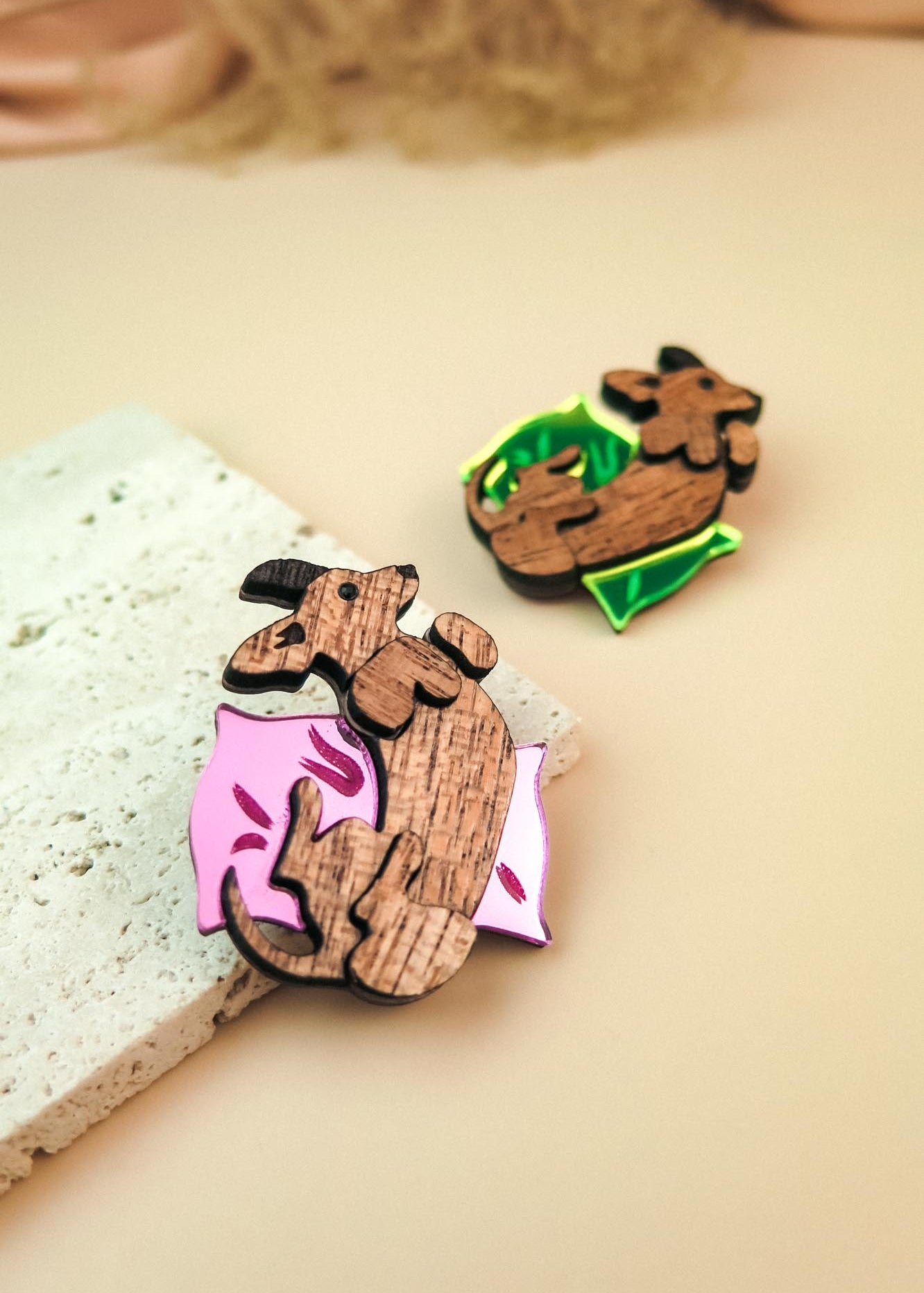 Two wooden pins shaped like dachshunds, one lying on a pink pillow with leaf designs and the other on a green pillow. Both are displayed as playful jewelry on a light beige surface next to a textured stone.