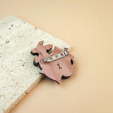 A pink, animal-shaped dog pin with a silver clasp lies on a textured, off-white stone surface against a soft beige background—perfect for lovers of playful jewelry.