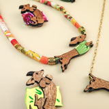 Playful jewelry pieces shaped like dachshund dogs are displayed on a neutral surface, including dachshund brooches and necklaces. Some feature colorful accents like green sweaters and floral designs, adding charm to each dog pin.