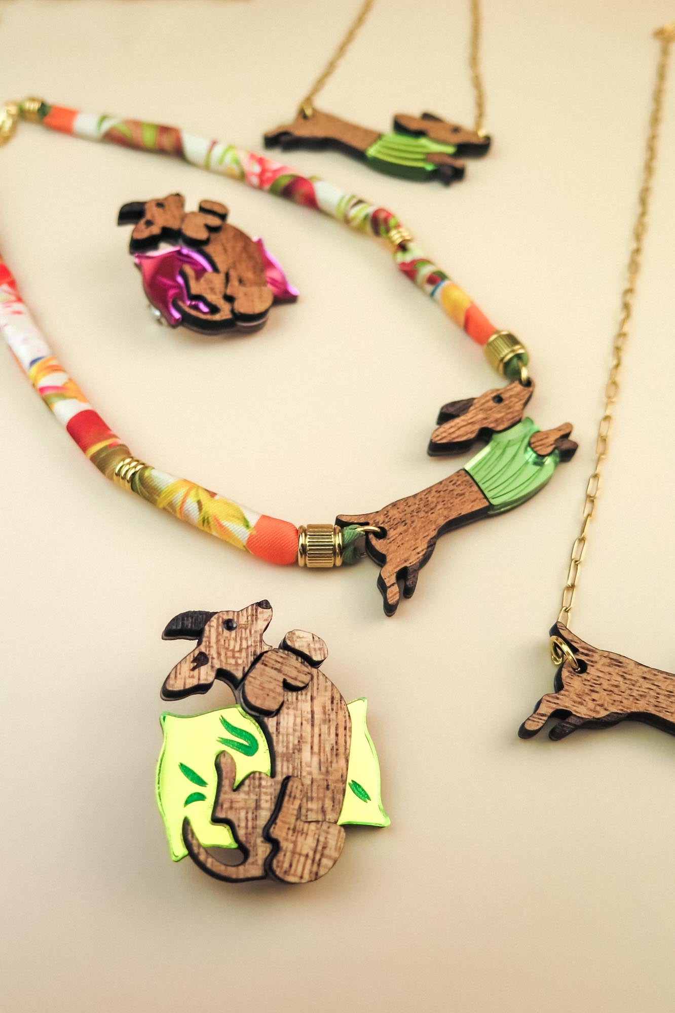 Playful jewelry pieces shaped like dachshund dogs are displayed on a neutral surface, including dachshund brooches and necklaces. Some feature colorful accents like green sweaters and floral designs, adding charm to each dog pin.