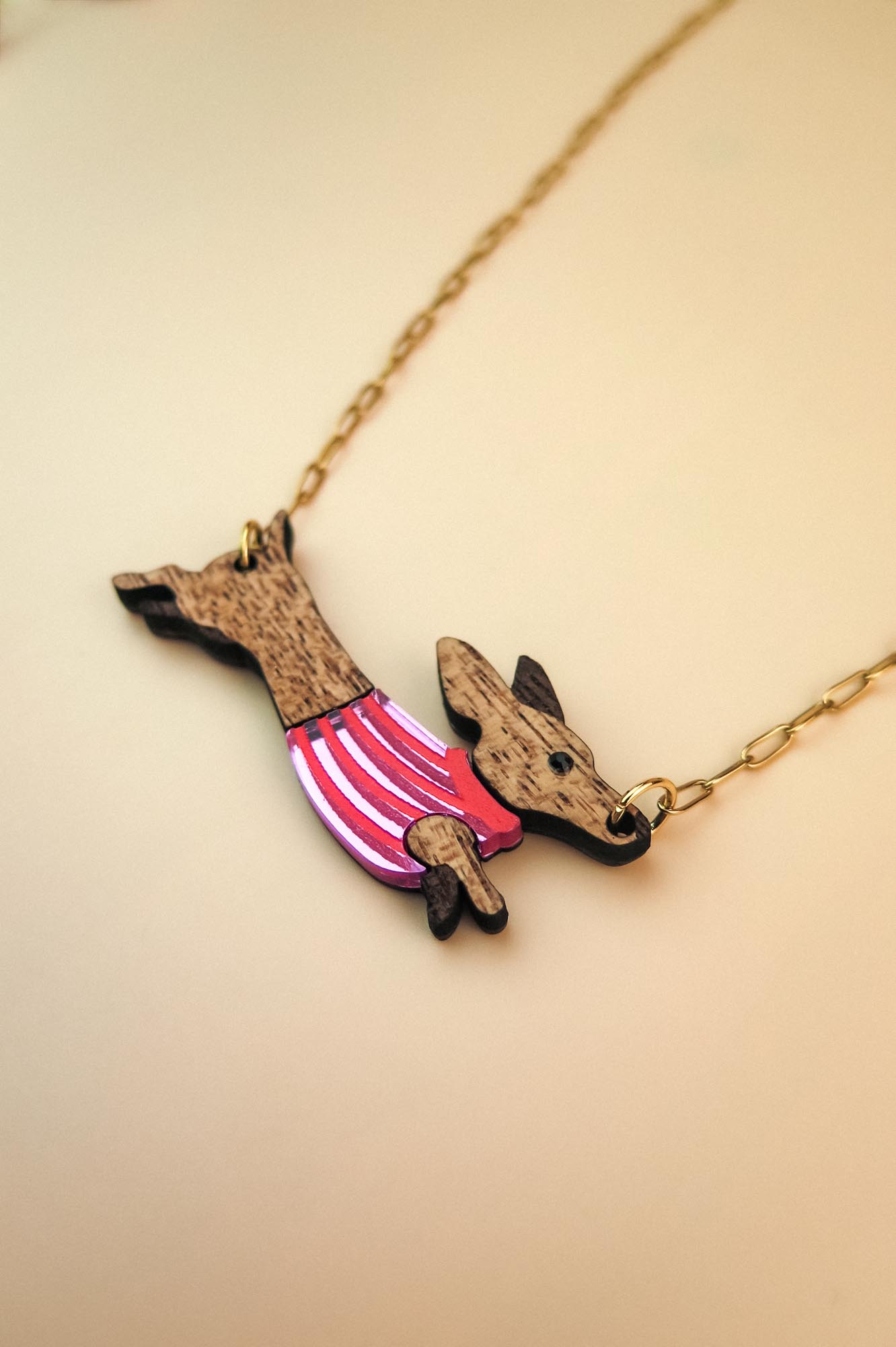 A necklace with a gold chain featuring a pendant shaped like a Dachshund in a colorful sweater, crafted from sustainable wood, set against a light beige background.