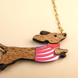 A wooden necklace pendant shaped like a dachshund dog in a colorful sweater, crafted from sustainable wood and attached to a gold chain, shown on a light beige background.