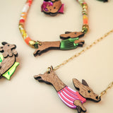 Colorful jewelry featuring Dachshund-shaped pendants crafted from sustainable wood, each dressed in a bright, colorful sweater. Displayed on a beige surface, the collection includes playful necklaces and pins with gold chains and vibrant beads.
