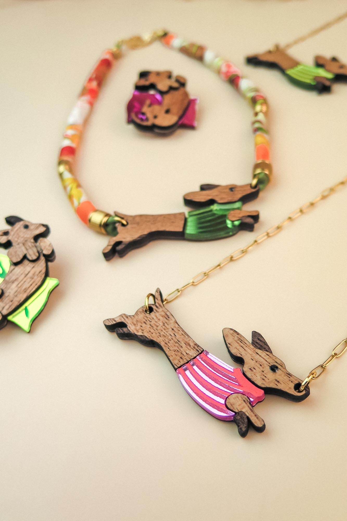Colorful jewelry featuring Dachshund-shaped pendants crafted from sustainable wood, each dressed in a bright, colorful sweater. Displayed on a beige surface, the collection includes playful necklaces and pins with gold chains and vibrant beads.