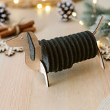 A napkin holder shaped like a dachshund dog, with dark gray napkins forming the body, sits on a light wooden table. Dachshund coasters and festive lights with pine cones are blurred in the background.