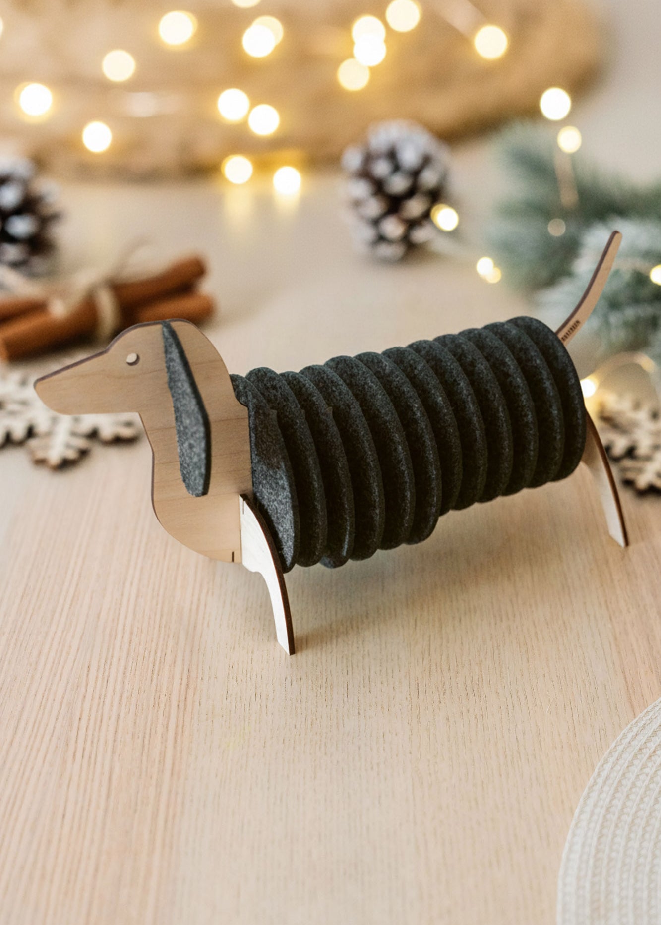 A napkin holder shaped like a dachshund dog, with dark gray napkins forming the body, sits on a light wooden table. Dachshund coasters and festive lights with pine cones are blurred in the background.