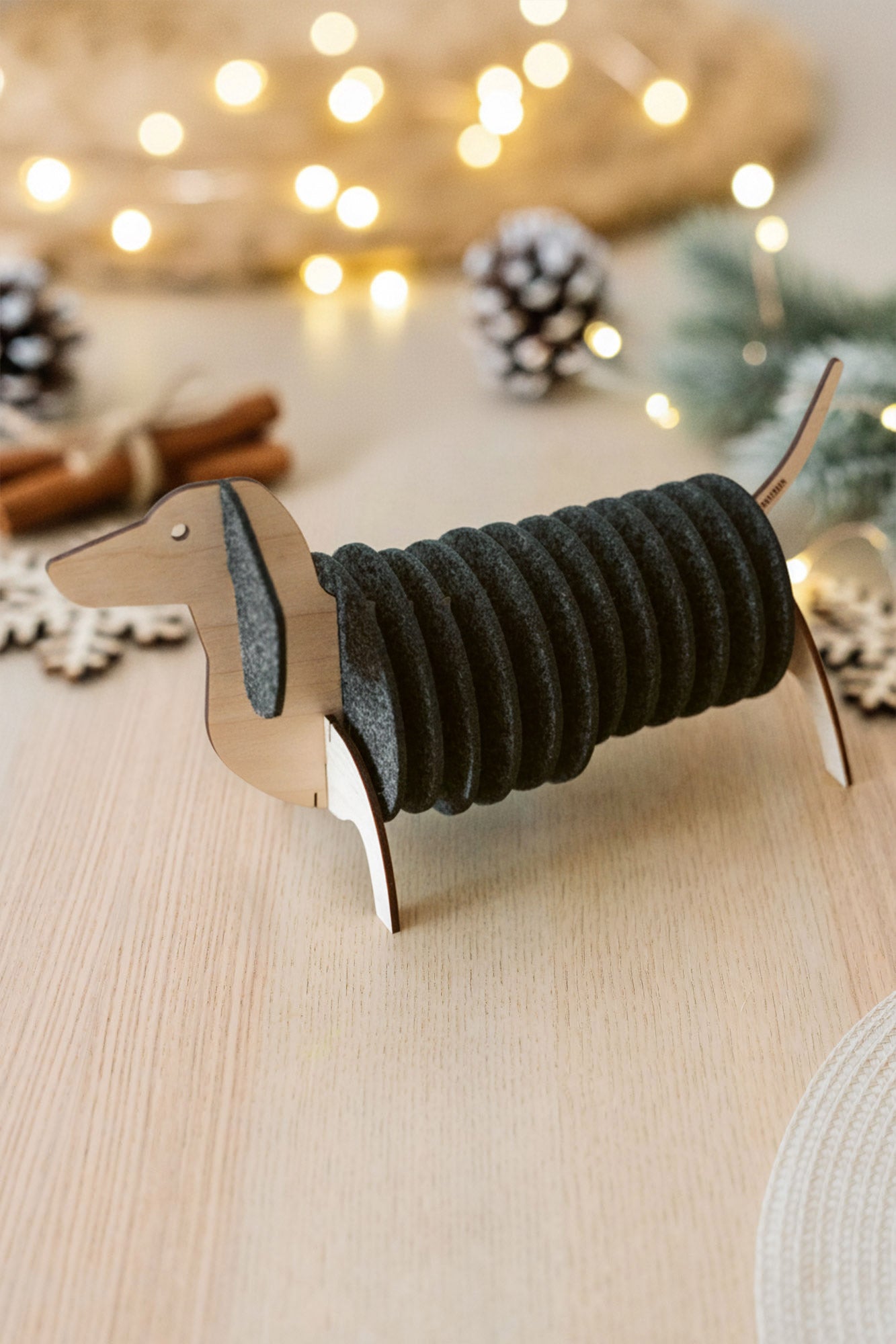 A napkin holder shaped like a dachshund dog, with dark gray napkins forming the body, sits on a light wooden table. Dachshund coasters and festive lights with pine cones are blurred in the background.