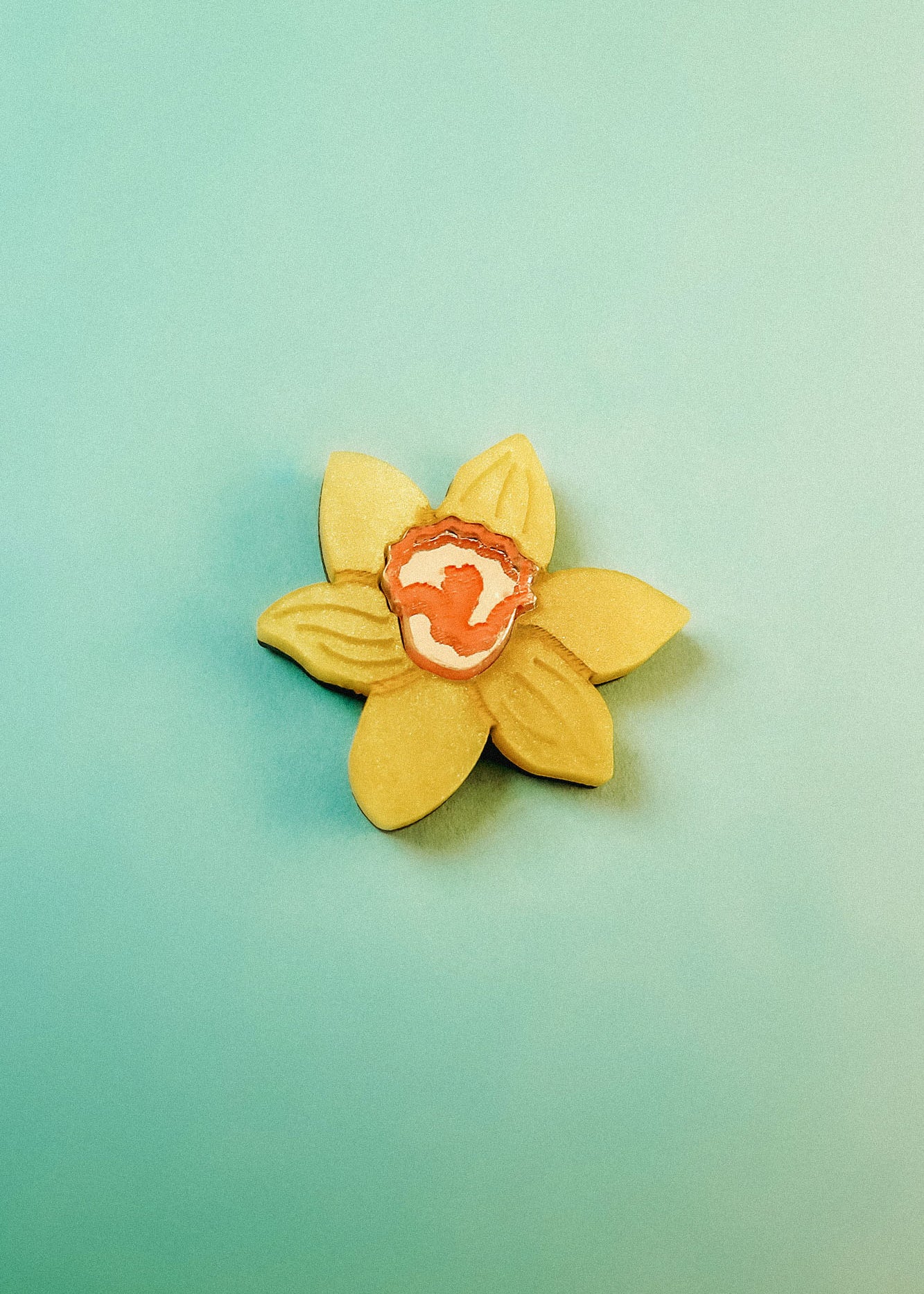 A handmade yellow flower-shaped cookie with an orange and white center is placed on a light teal background, reminiscent of a Daffodil Flower Lapel Pin crafted in acrylic.