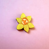 A handmade yellow Daffodil Flower Lapel Pin with an orange center is placed on a pink and purple gradient background.