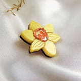 A handmade yellow Daffodil Flower Lapel Pin brooch with a small orange and white cameo in the center, crafted from acrylic, rests on soft white satin fabric. Delicate dried plant stems add a subtle touch in the background.