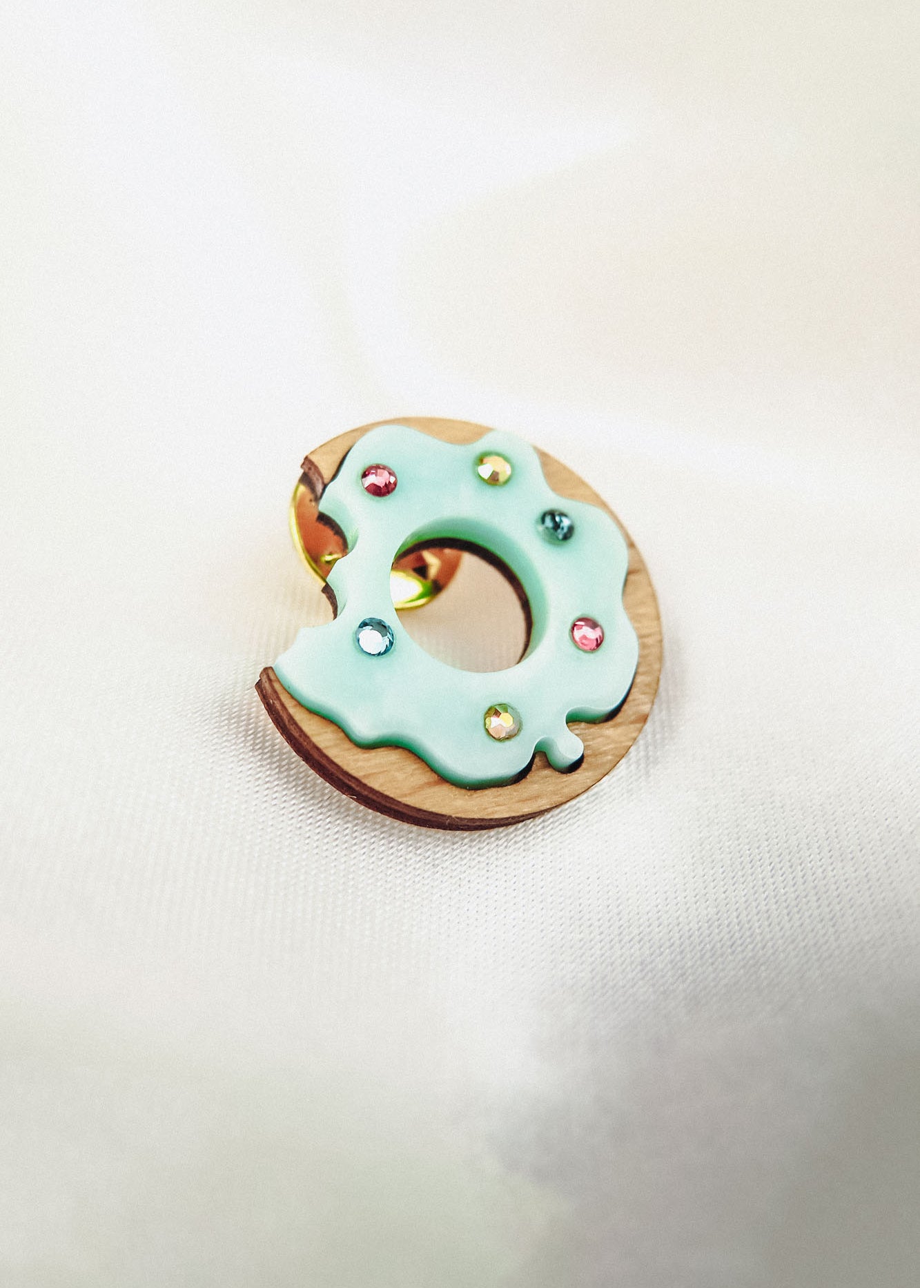 A handcrafted wooden Donut Pin shaped like a bitten donut, topped with pale blue icing and colorful sprinkles, rests on a soft white fabric background.